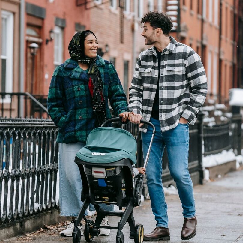 Full body of multiracial parents looking at each other while strolling on sidewalk along buildings with anonymous child in baby carriage